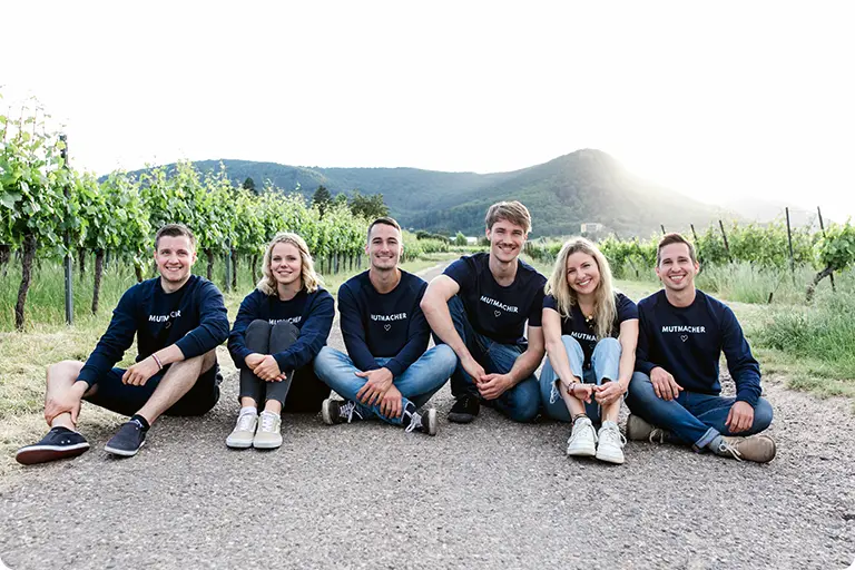 "Gruppe glücklicher Menschen in Freizeitkleidung, lächelnd und die Natur genießend, umgeben von einer malerischen Landschaft mit Bergen, Pflanzen und einem blauen Himmel."